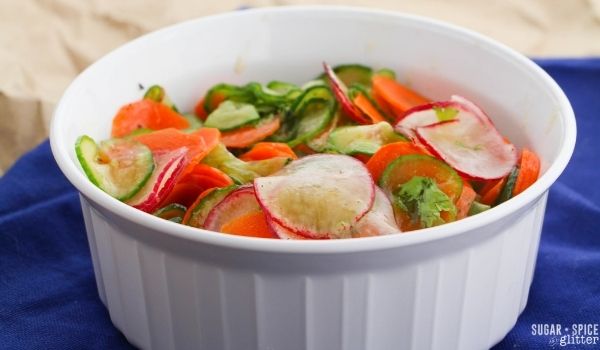 white bowl on a blue napkin filled with thinly sliced carrots, cucumber and radishes dressed with a cilantro-lime dressing