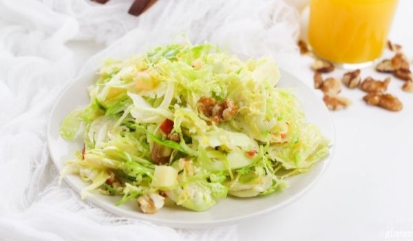 shaved Brussels sprouts salad with apples, walnuts, Parmesan and bacon on a white plate with orange-dijon salad dressing in the background