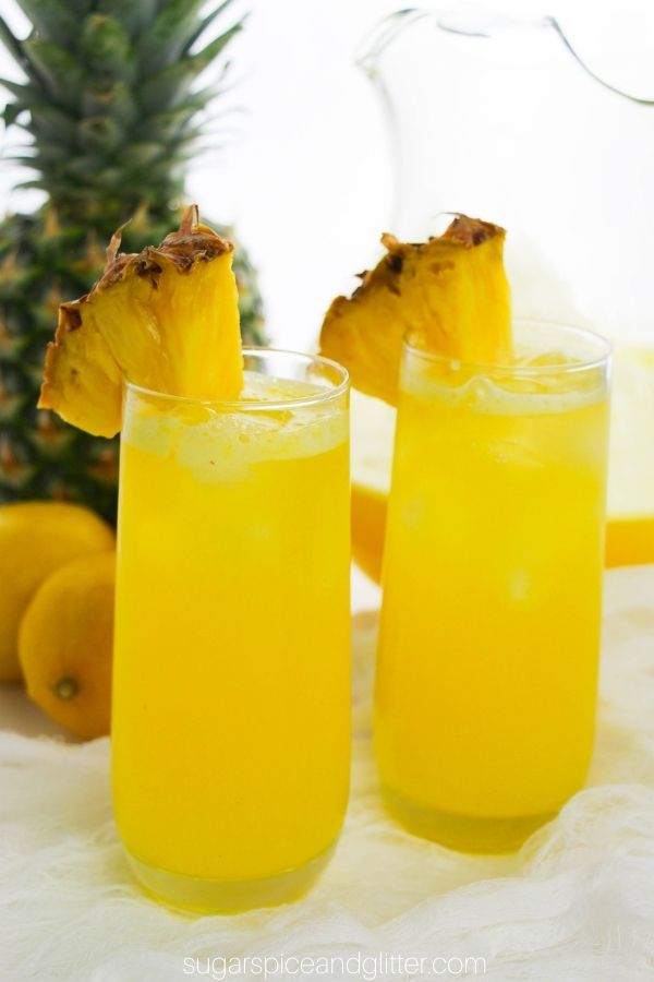 close-up image of two glasses of pineapple lemonade garnished with pineapple wedges, with a pineapple, lemons and a pitcher of the lemonade in the background