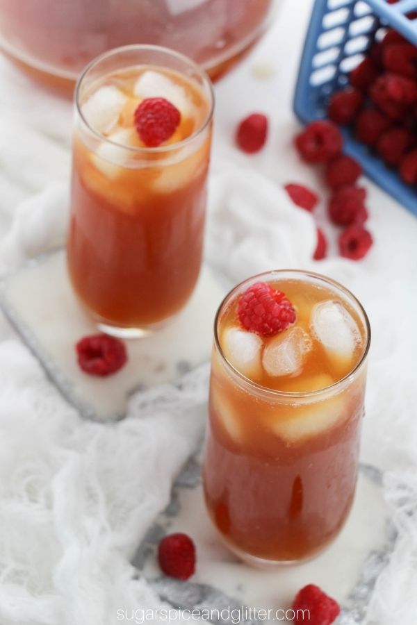 overhead shot of two tall glasses filled with rasperry iced tea, garnished with raspberries with a pitcher of raspberry iced tea and a carton of raspberries in the background