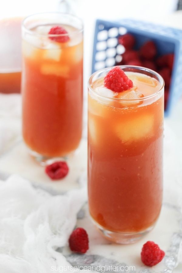 two tall glasses filled with rasperry iced tea, garnished with raspberries with a pitcher of raspberry iced tea and a carton of raspberries in the background