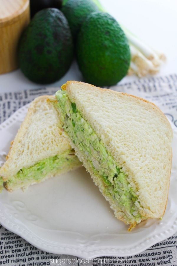 a sliced avocado chicken sandwich on a white plate with ingredients needed to make it in the background