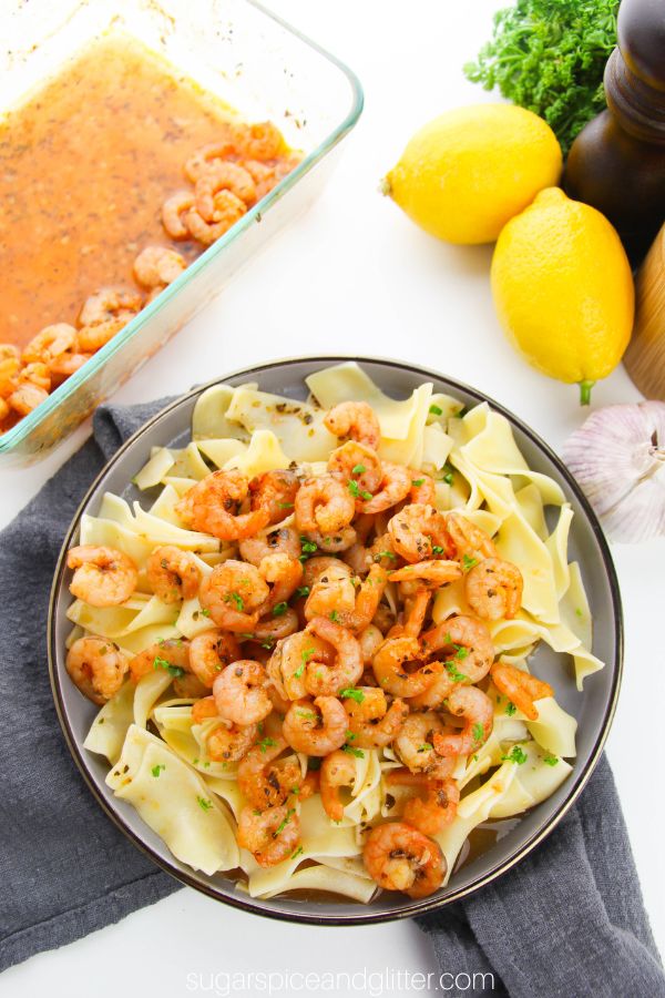 overhead image of a gray plate piled with egg noodles and plenty of baked lemon garlic shrimp, along with the ingredients needed to make the shrimp in the background