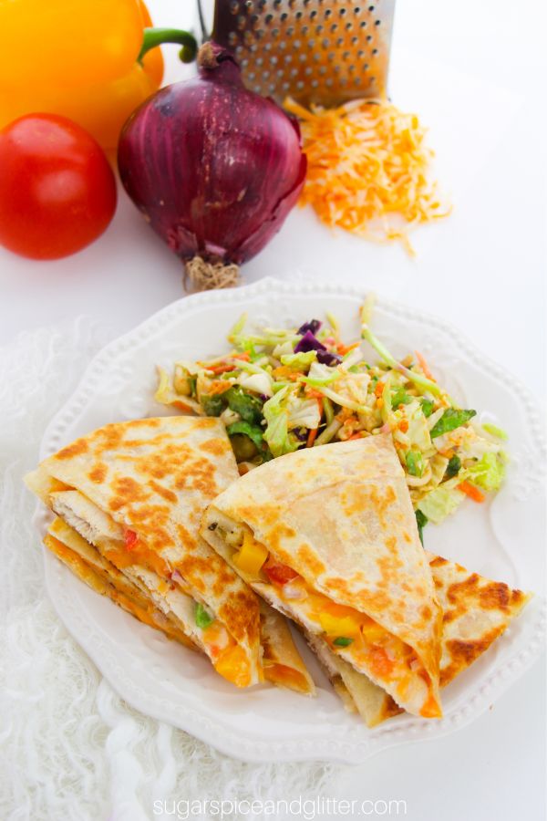 overhead shot of a white plate filled with chicken quesadillas and a side salad, with ingredients to make a chicken quesadilla in the background