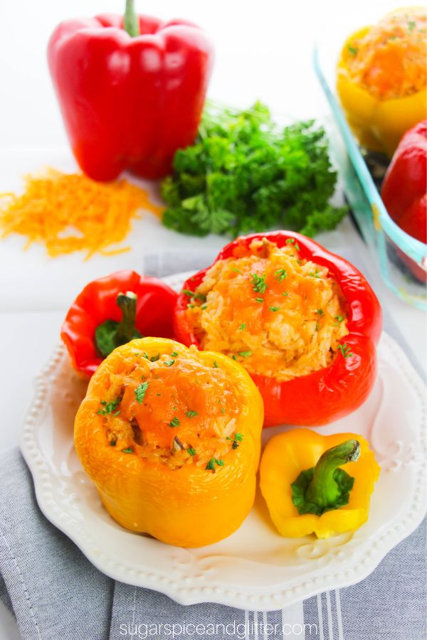 bright bell peppers stuffed with cheese, rice and buffalo chicken on a white plate with cheese, more peppers and parsley in the background