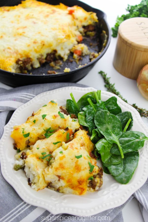 a white plate with a generous helping of cottage pie served alongside a simple spinach salad with a cast-iron skillet full of cottage pie in the background