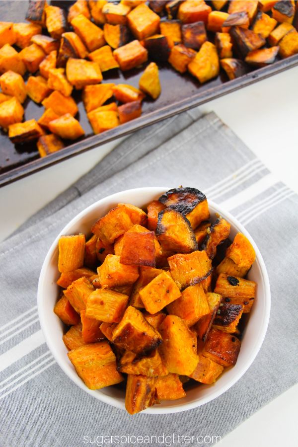 overhead image of a white bowl full of bright orange roasted sweet potatoes along with a sheet pan of more roasted sweet potatoes in the background