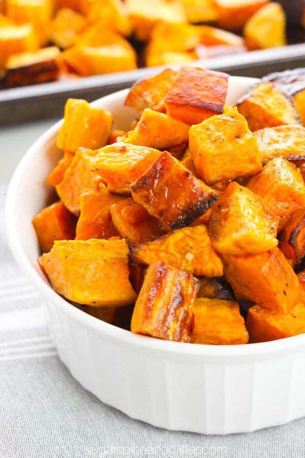 close-up image of a white bowl full of bright orange roasted sweet potatoes along with a sheet pan of more roasted sweet potatoes in the background