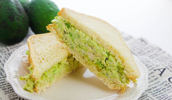 a sliced avocado chicken sandwich on a white plate with ingredients needed to make it in the background