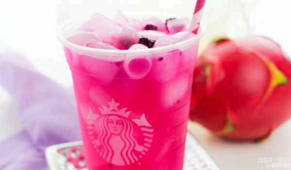 close-up image of a Starbucks cup filled with ice and pink dragonfruit lemonade on a silver coaster on top of a purple napkin, with a dragonfruit in the background