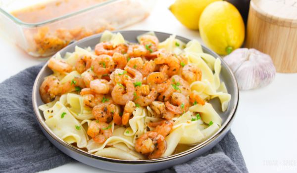 image of a gray plate piled with egg noodles and plenty of baked lemon garlic shrimp, along with the ingredients needed to make the shrimp in the background