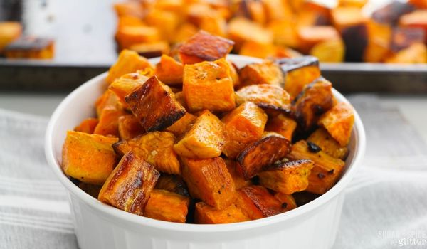 white bowl full of bright orange roasted sweet potatoes along with a sheet pan of more roasted sweet potatoes in the background