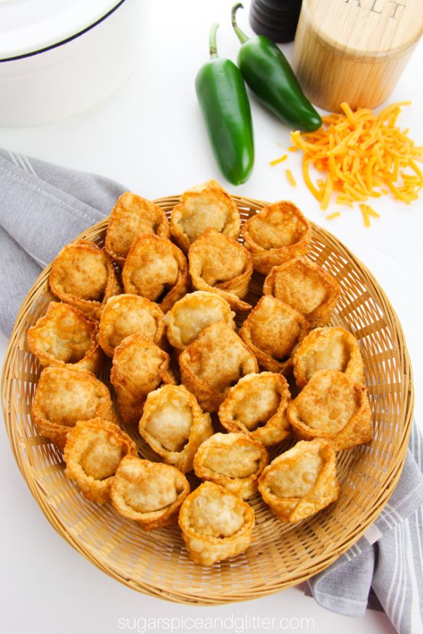 overhead picture of a wicker basket filled with jalapeno popper wontons on a gray napkin with jalapenos, cheddar cheese and seasonings in the background