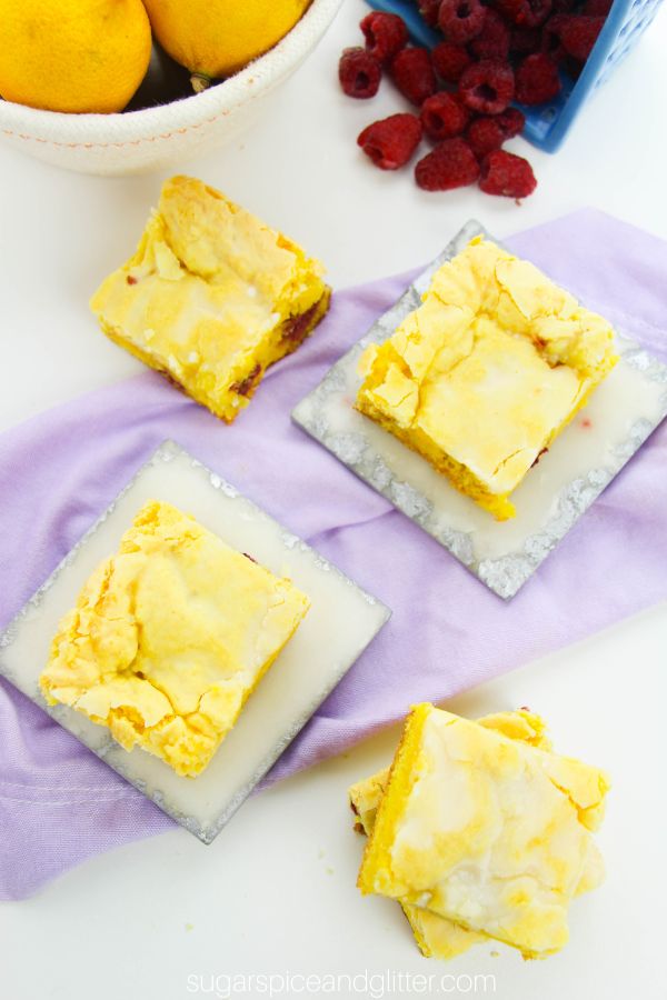 overhead image of a batch of lemon raspberry blondies on white and silver coasters and a lilac napkin with lemons and raspberries in the background
