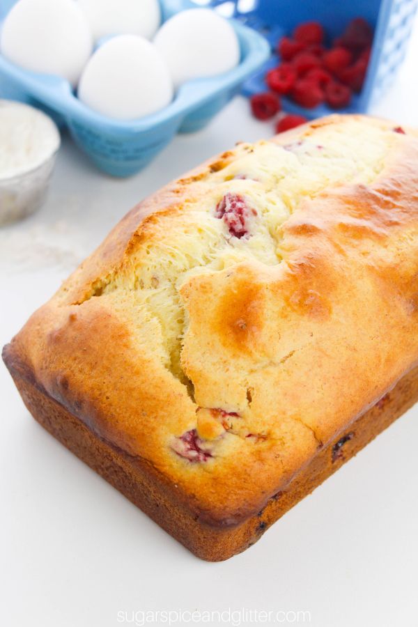 close-up image of a lemon raspberry loaf cake with the ingredients needed to make it in the background
