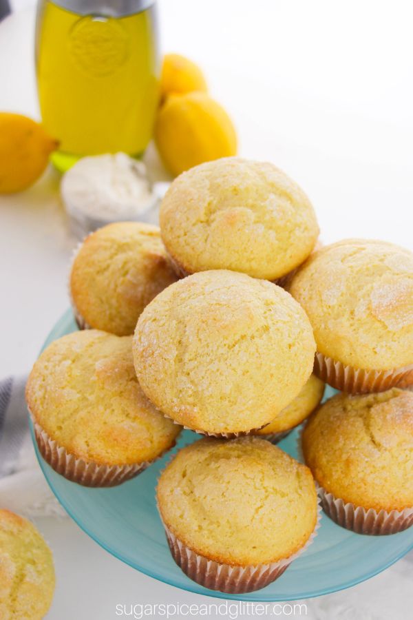 overhead image of lemon ricotta muffins with a light sugar dusting on a blue cake pedestal with lemons, olive oil and ricotta cheese in the background