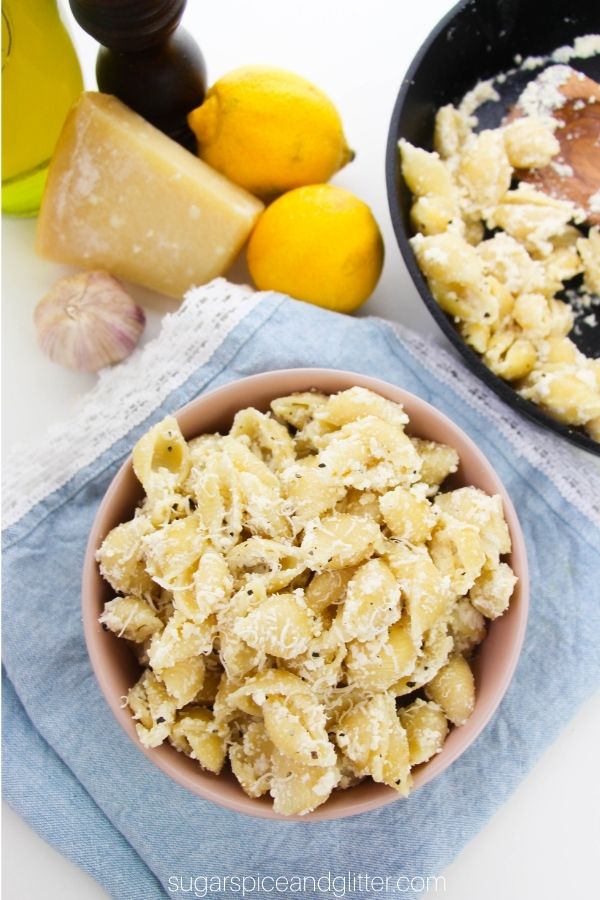 a pink bowl filled with creamy Lemon Ricotta pasta set on a denim napkin with a cast-iron pan holding more of the pasta in the background, along with ingredients needed to make the recipe