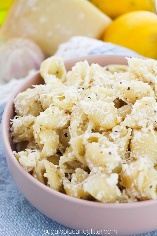 a pink bowl filled with creamy Lemon Ricotta pasta set on a denim napkin with the ingredients needed to make the recipe in the background