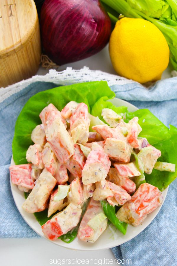 overhead image of creamy, cold crab salad on a bed of lettuce on a white plate set on top of a blue napkin with red onion, lemon and celery in the background