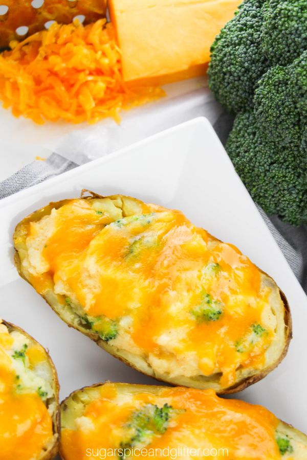 cheesy twice baked potatoes with bits of steamed broccoli peeking out from under the cheese, set on a white plate, with cheese and broccoli in the background