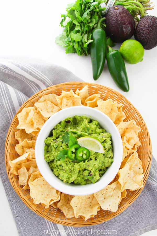 a white bowl filled with guacamole, garnished with jalapenos, lime and cilantro, set on a wicker tray holding tortilla chips with ingredients needed to make the guacamole in the background