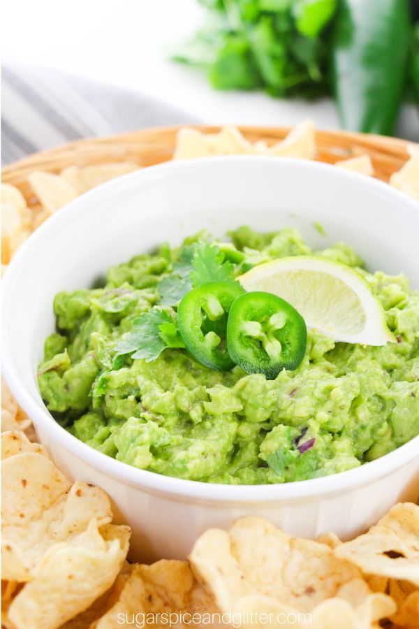 a white bowl filled with guacamole, garnished with jalapenos, lime and cilantro, set on a wicker tray holding tortilla chips