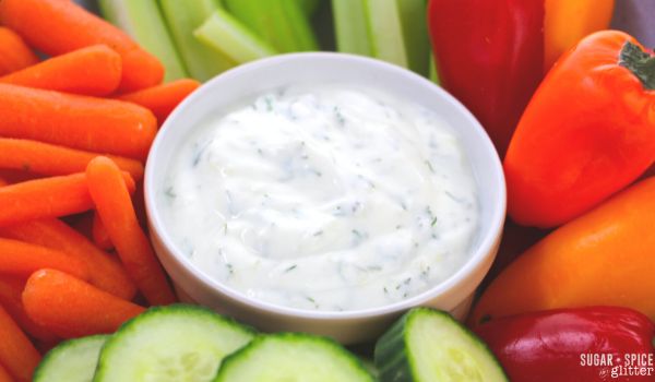 a beige bowl holding dill veggie dip, surrounded by sliced veggies
