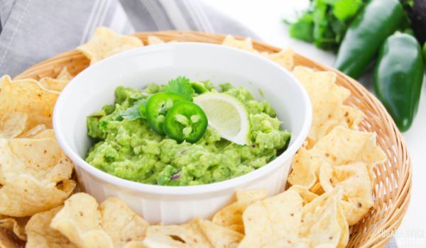 a white bowl filled with guacamole, garnished with jalapenos, lime and cilantro, set on a wicker tray holding tortilla chips