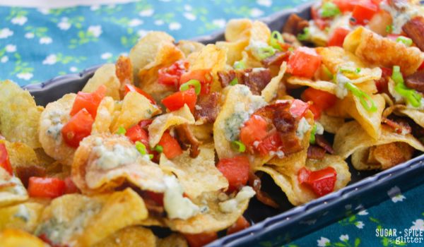close-up image of kettle chips on a blue platter topped with melted blue cheese, tomatoes, bacon and green onions