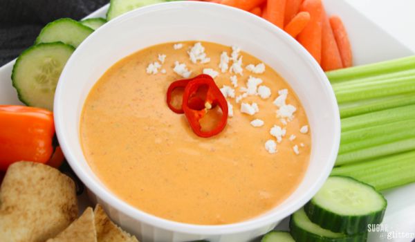 a white bowl filled with roasted red pepper dip, garnished with sliced red peppers and a sprinkle of feta cheese