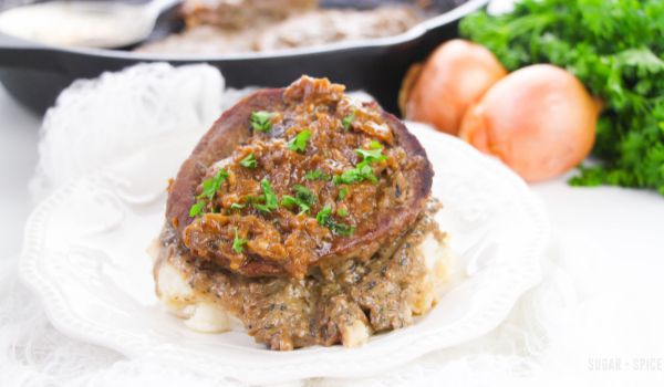 steak smothered in onion gravy and sprinkled with parsley on a white plate with a cast-iron pan in the background