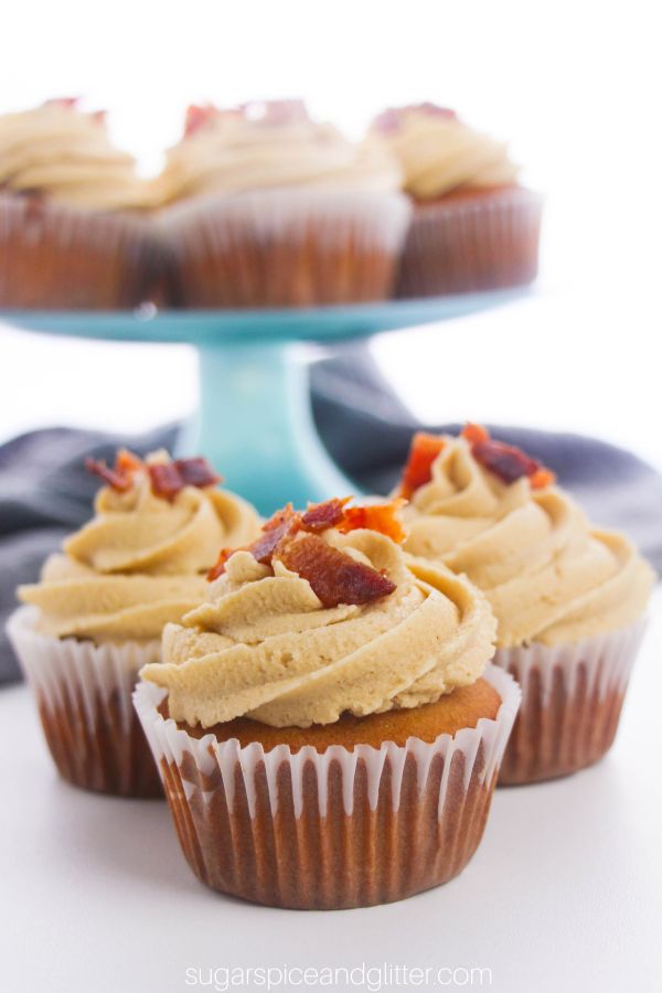 a trio of maple bacon cupcakes with more cupcakes in the background on a blue cake pedestal