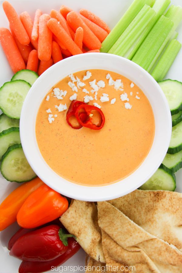 a white bowl filled with roasted red pepper dip, garnished with sliced red peppers and a sprinkle of feta cheese set on a platter with veggies and pita bread