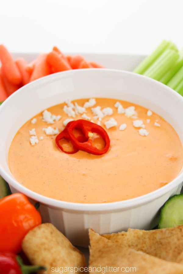 a white bowl filled with roasted red pepper dip, garnished with sliced red peppers and a sprinkle of feta cheese set on a platter with veggies and pita bread
