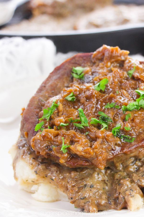 close-up of a steak smothered in onion gravy and sprinkled with parsley on a white plate with a cast-iron pan in the background