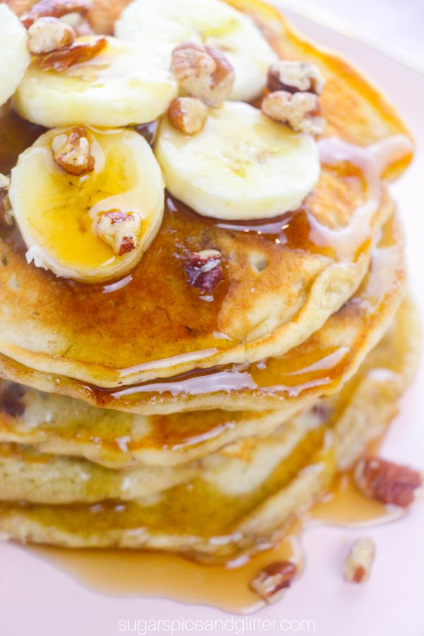 a stack of banana nut pancakes topped with sliced bananas, chopped pecan and drizzled with plenty of maple syrup on a pink plate