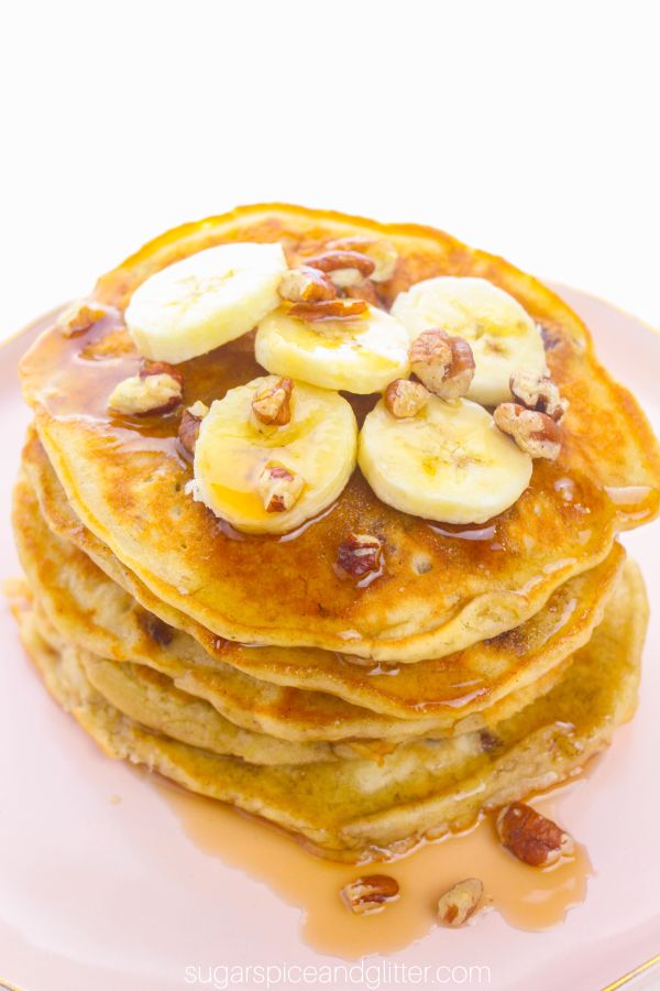 a stack of banana nut pancakes topped with sliced bananas, chopped pecan and drizzled with plenty of maple syrup on a pink plate