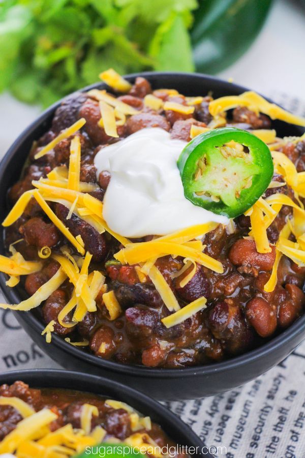 overhead image of a black bowl filled with beef and bean chili topped with sour cream, shredded cheddar cheese and a jalapeno pepper slice