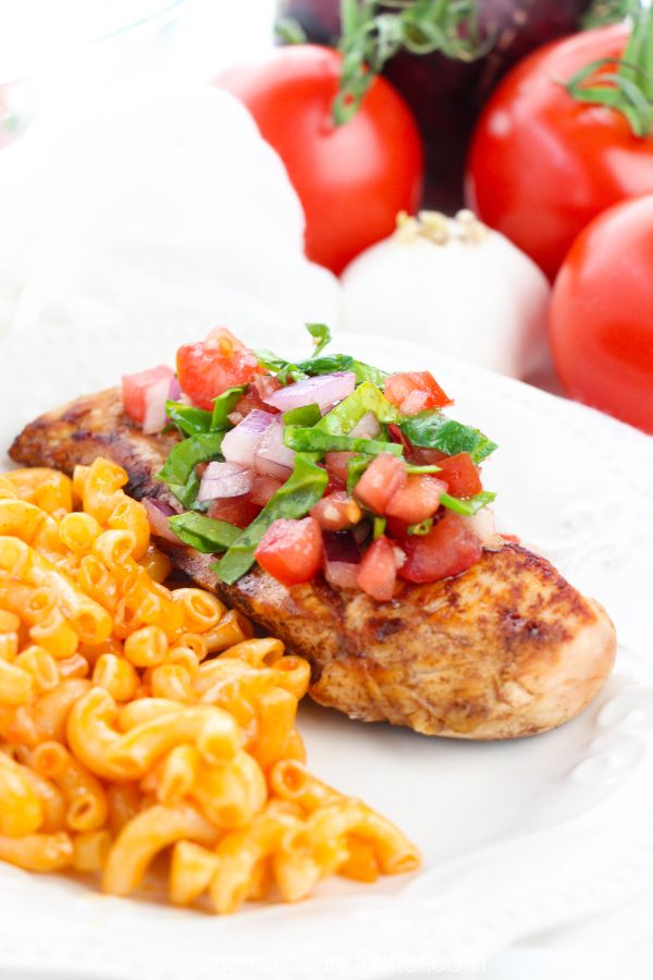 close-up shot of a white plate with a marinated chicken breast topped with a bruschetta tomato mixture, along with a pasta salad and the ingredients for the bruschetta chicken in the background