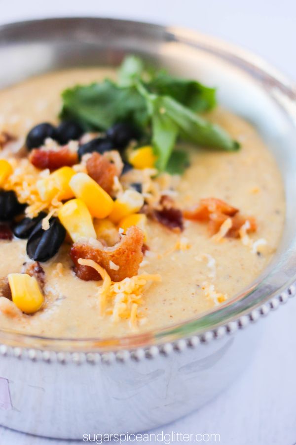 close-up image of corn chowder in a silver bowl with corn, black beans, bacon, cilantro and cheese sprinkled on top
