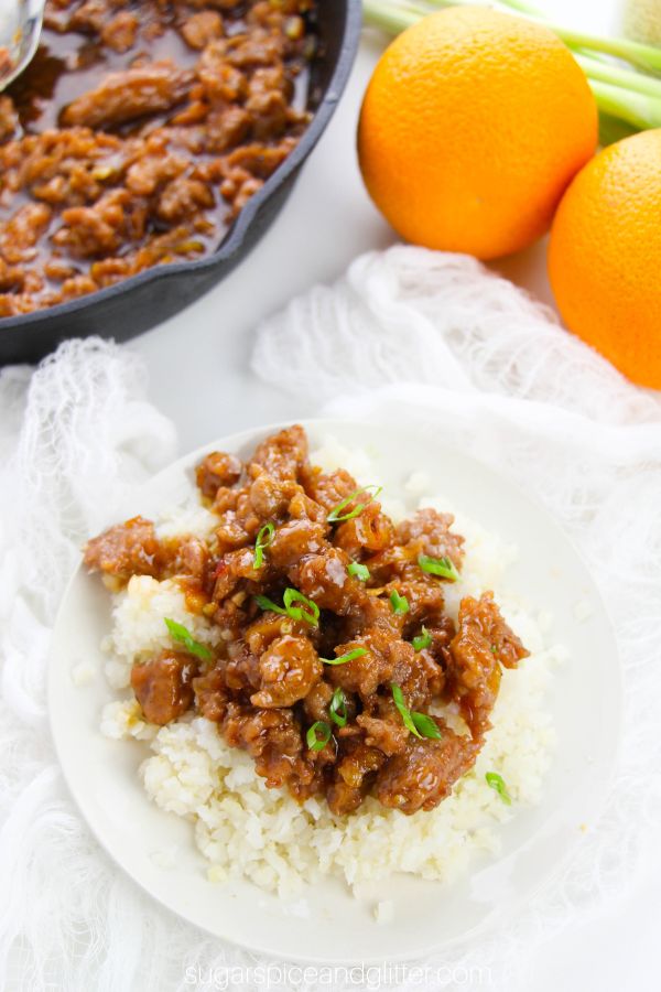 Saucey orange chicken on a bed of white rice on a white plate sprinkled with green onions, with a skillet of orange chicken and two oranges and more green onions in he background
