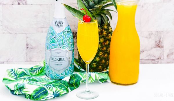 picture of a champagne flute filled with a pineapple mimosa, topped with a pineapple leaf and cherry garnish, with the ingredients needed to make the cocktail in the background