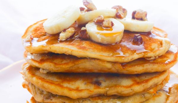 a stack of banana nut pancakes topped with sliced bananas, chopped pecan and drizzled with plenty of maple syrup