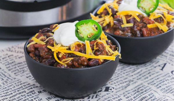 two black bowls filled with beef and bean chili topped with sour cream, shredded cheddar cheese and a jalapeno pepper slice on a newsprint napkin