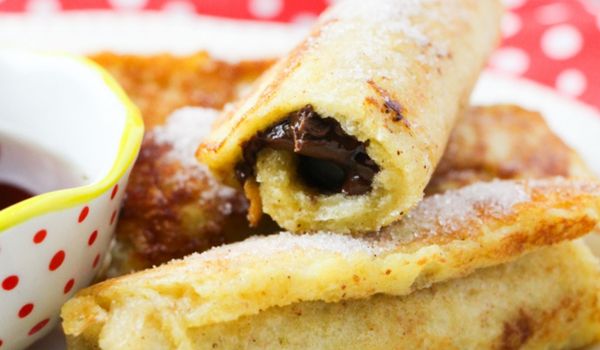 close-up image of a stack of nutella french toast rolls, showing the inside filling, on a white plate with a small bowl of maple syrup