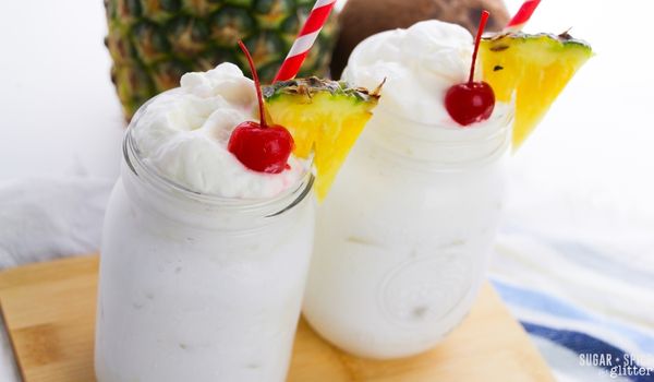 two mason jars filled with whipped pina colada cocktails topped with pineapple whipped cream and garnished with maraschino cherries and pineapple wedges on a wooden tray with a pineapple and coconut in the background