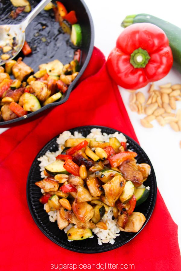 Kung Pao chicken on a bed of rice on a black circular plate set on a red napkin with a skillet of more stirfry in the background