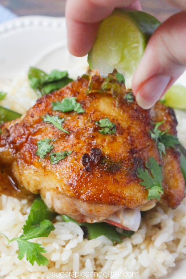 a golden lime cilantro chicken thigh on a bed of rice on a whilte plate with a hand squeezing a wedge of lime above the chicken