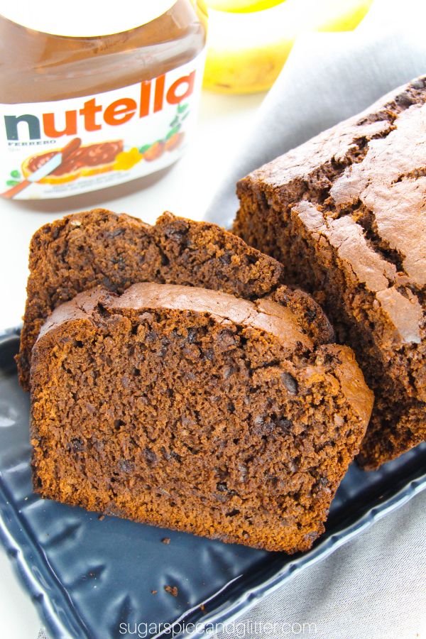 Nutella Banana Bread on a navy blue platter with two slices cut out to show the mini chocolate chips speckled throughout, with ingredients needed to make the recipe in the background