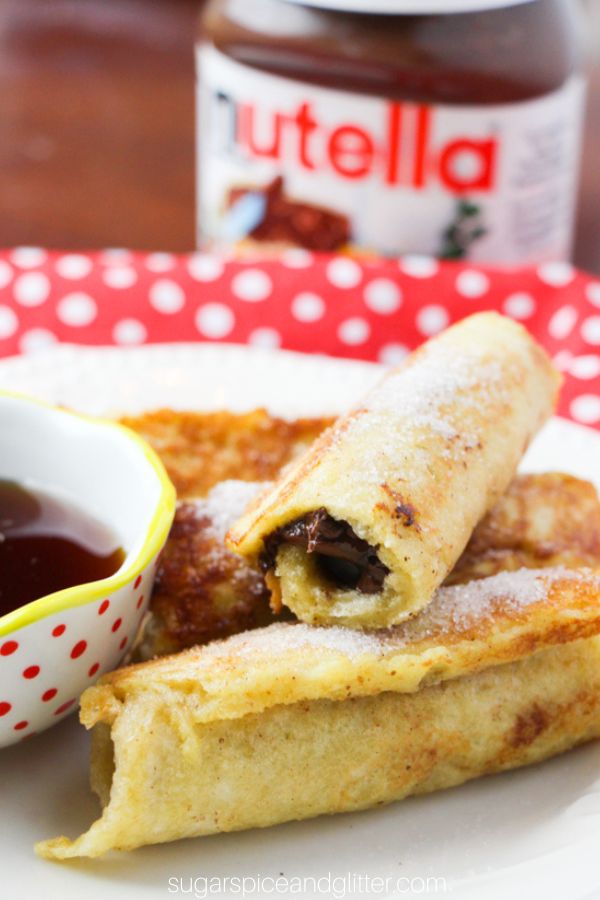close-up image of a stack of nutella french toast rolls, showing the inside filling, on a white plate with a small bowl of maple syrup and a jar of Nutella in the background